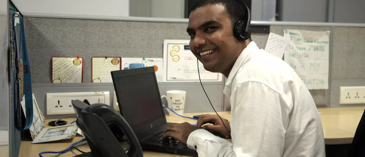 An Indian man smiles and looks at the camera while seated at an office cubicle.